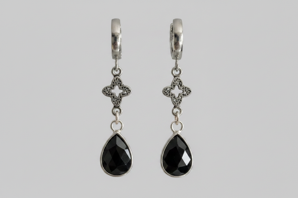 A pair of silver huggie hoop earrings with ornate filigree clover charms and faceted black teardrop gemstones, shown against a solid gray background.