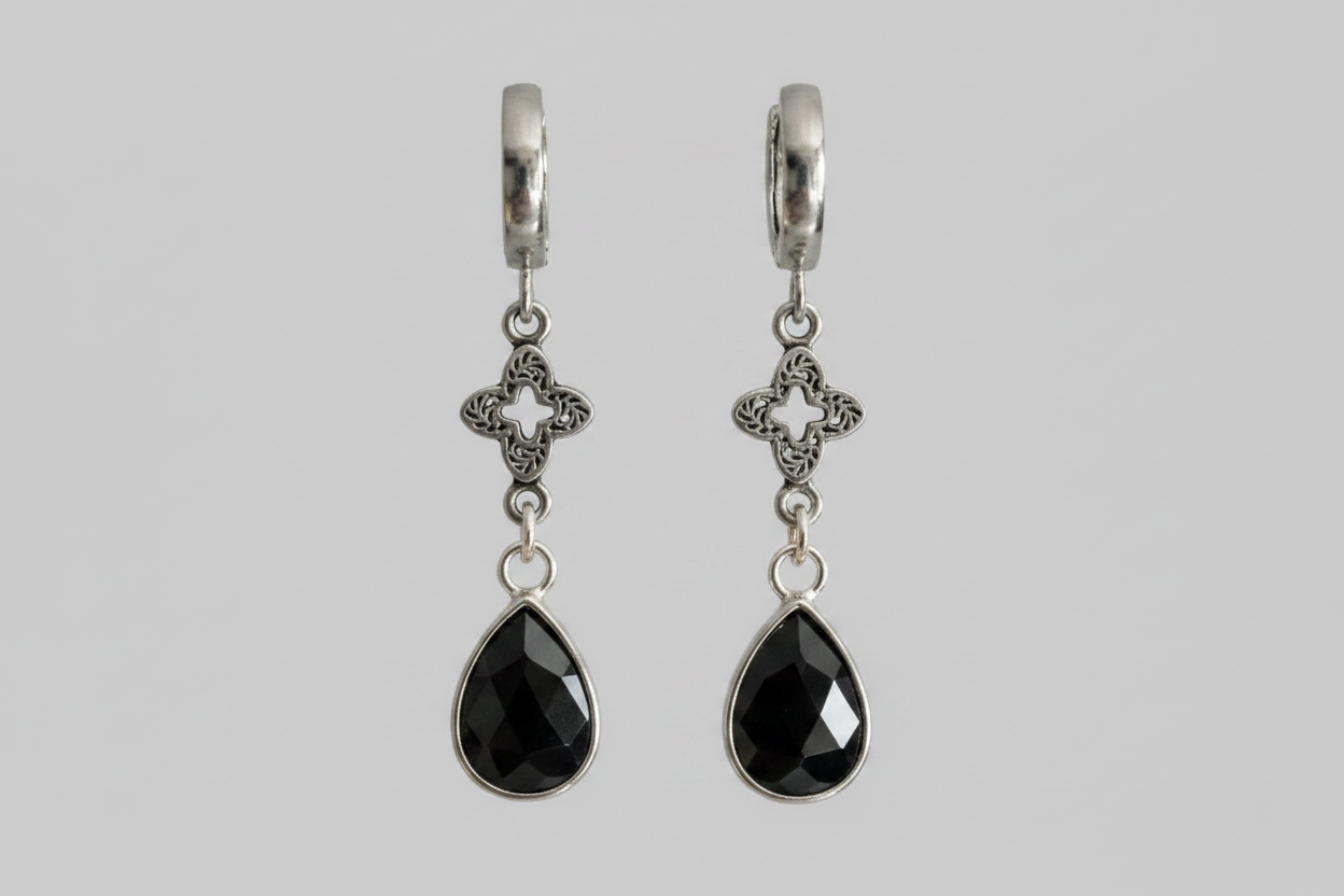 A pair of silver huggie hoop earrings with ornate filigree clover charms and faceted black teardrop gemstones, shown against a solid gray background.
