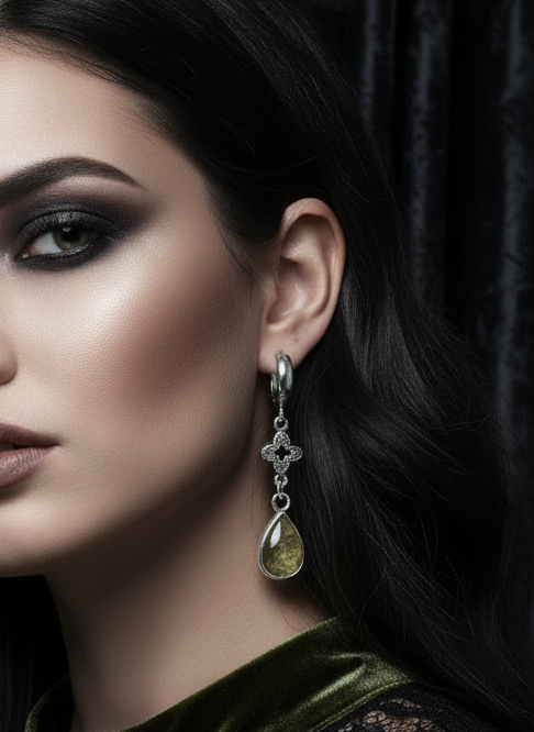 A close-up portrait of a woman with dark hair wearing silver dangle earrings with teardrop-shaped olive green gemstones and ornate silver filigree details, set against a dark draped background.