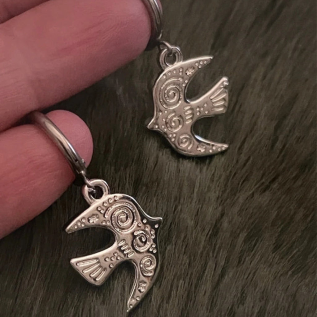 Silver bird-shaped earrings held between fingers on a dark textured surface