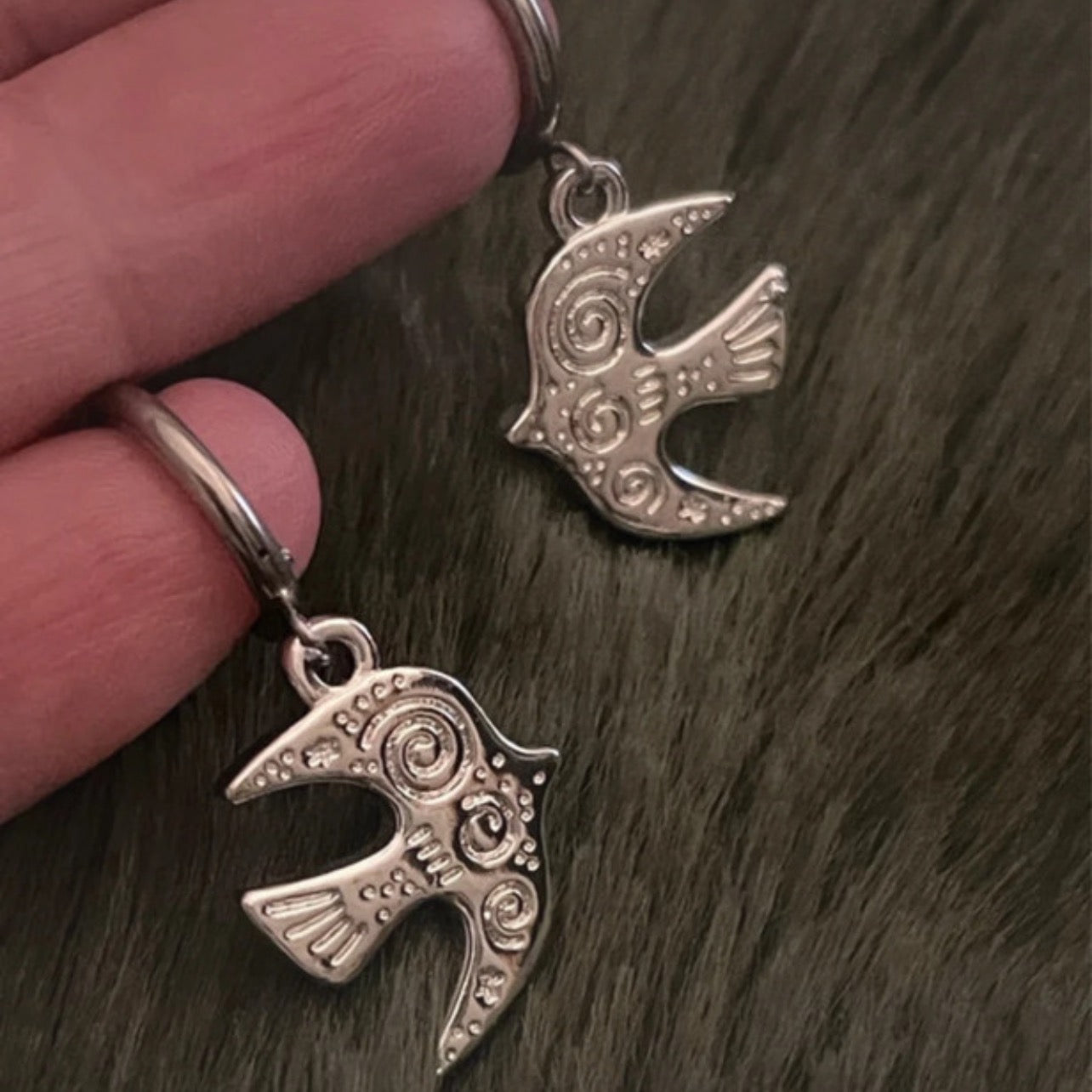 Silver bird-shaped earrings held between fingers on a dark textured surface