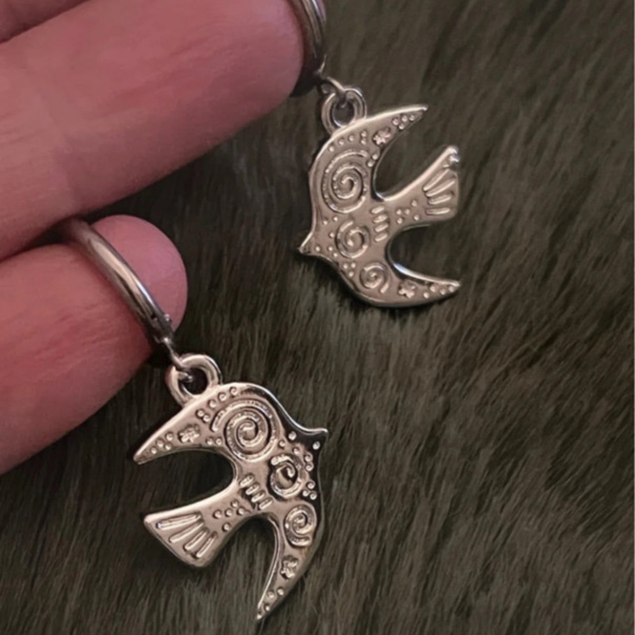 Silver bird-shaped earrings held by a hand on a dark textured surface