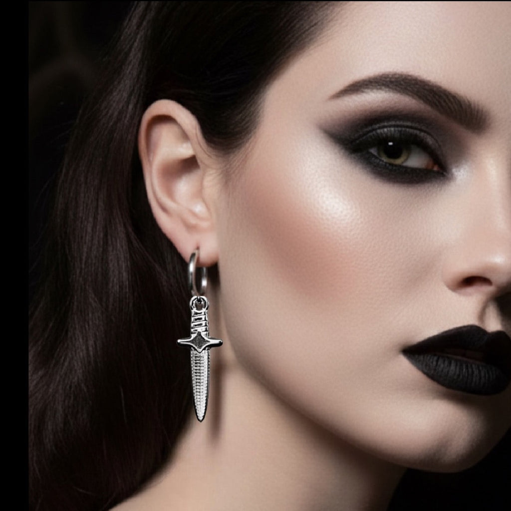 A close-up of a model's ear wearing a silver hoop earring with a dangling dagger-shaped charm, set against a dark background.