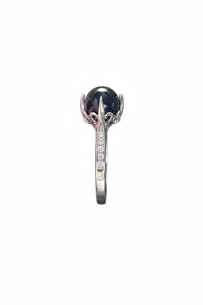 A silver-toned ring featuring a large, round, dark black gemstone in a prong setting, with a band decorated with small clear stones, shown against a plain white background.
