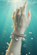 A close-up of a person's hand and wrist underwater, wearing a silver beaded bracelet with a crescent moon charm and a small round charm, surrounded by bubbles and light rays filtering through the water.