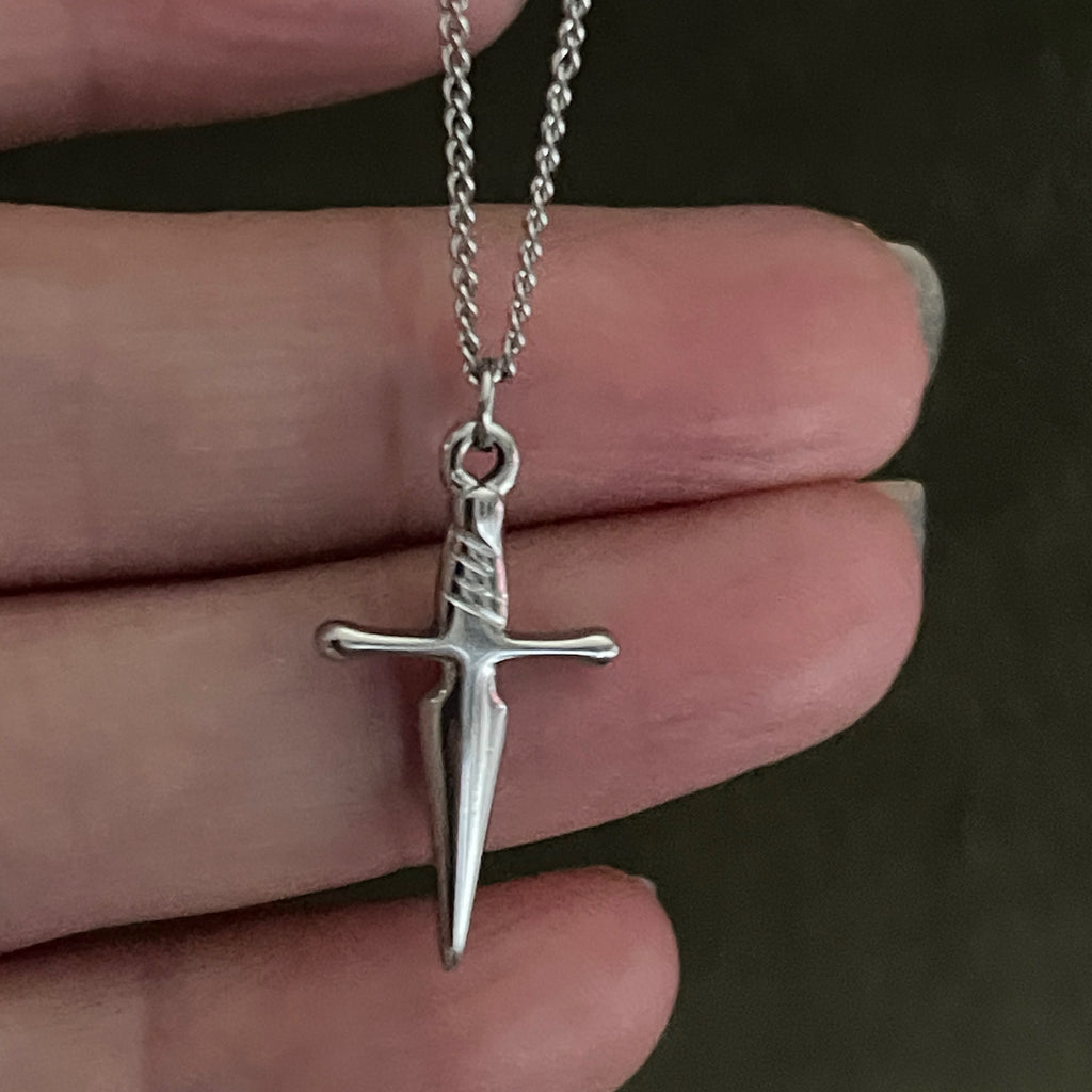 Silver sword-shaped pendant on a chain held between fingers against a dark background