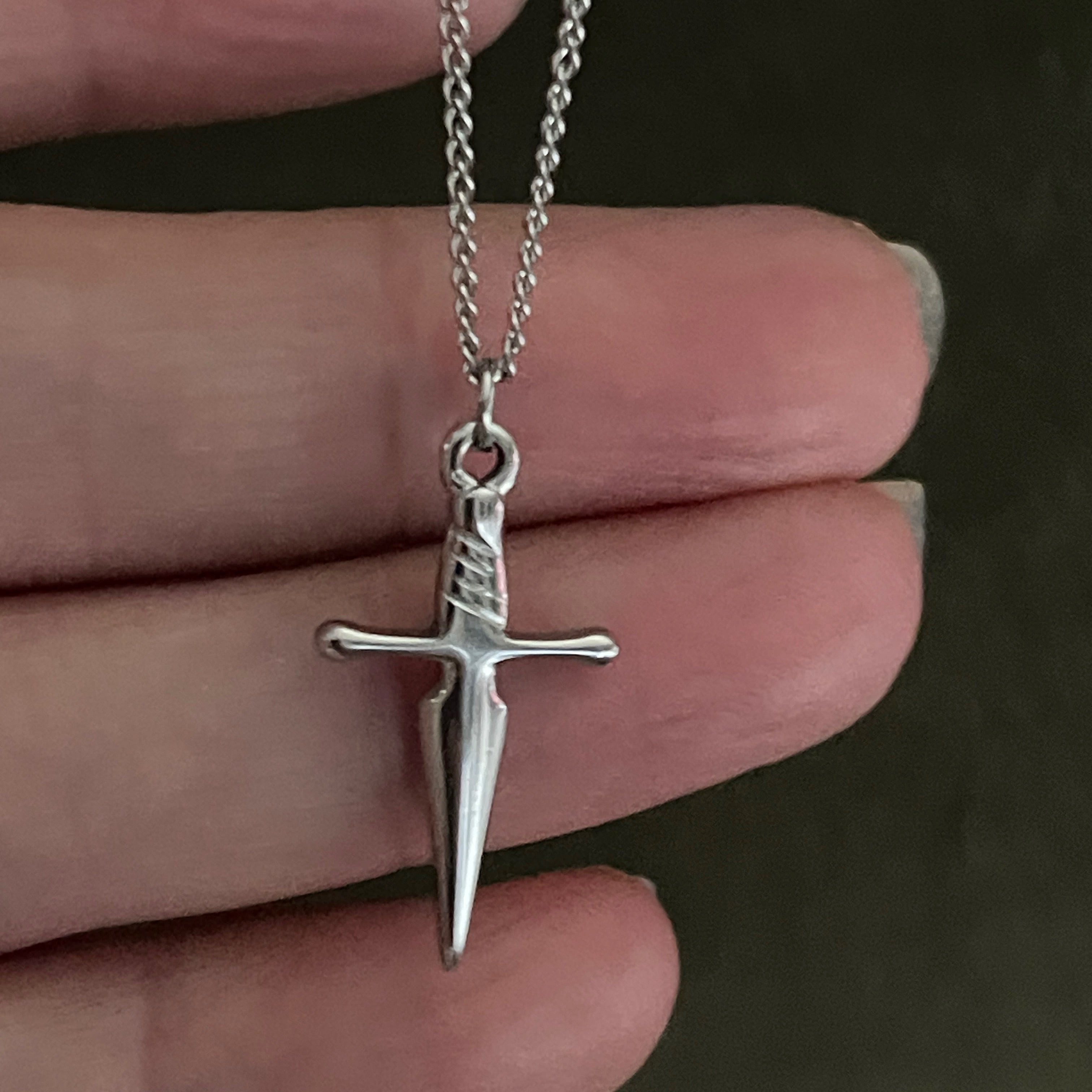 Silver sword-shaped pendant on a chain held between fingers against a dark background