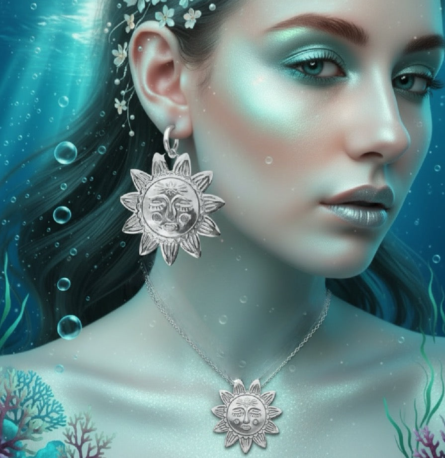 A close-up portrait of a woman underwater wearing a silver sun-face pendant necklace and matching dangling earring. The scene is blue-toned with bubbles, water, and coral-like marine details in the background.