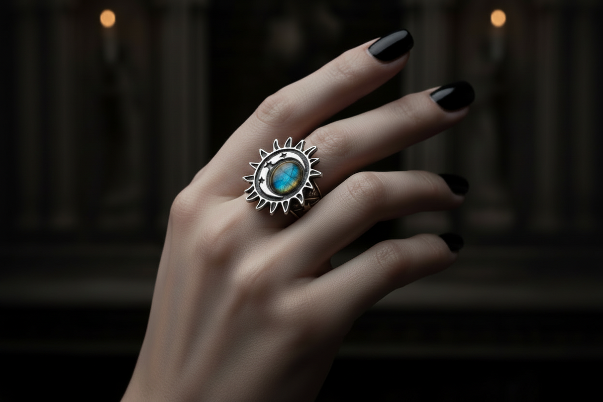 A hand with black nail polish wearing a silver sunburst ring featuring a blue and green iridescent stone, set against a dark, atmospheric background.