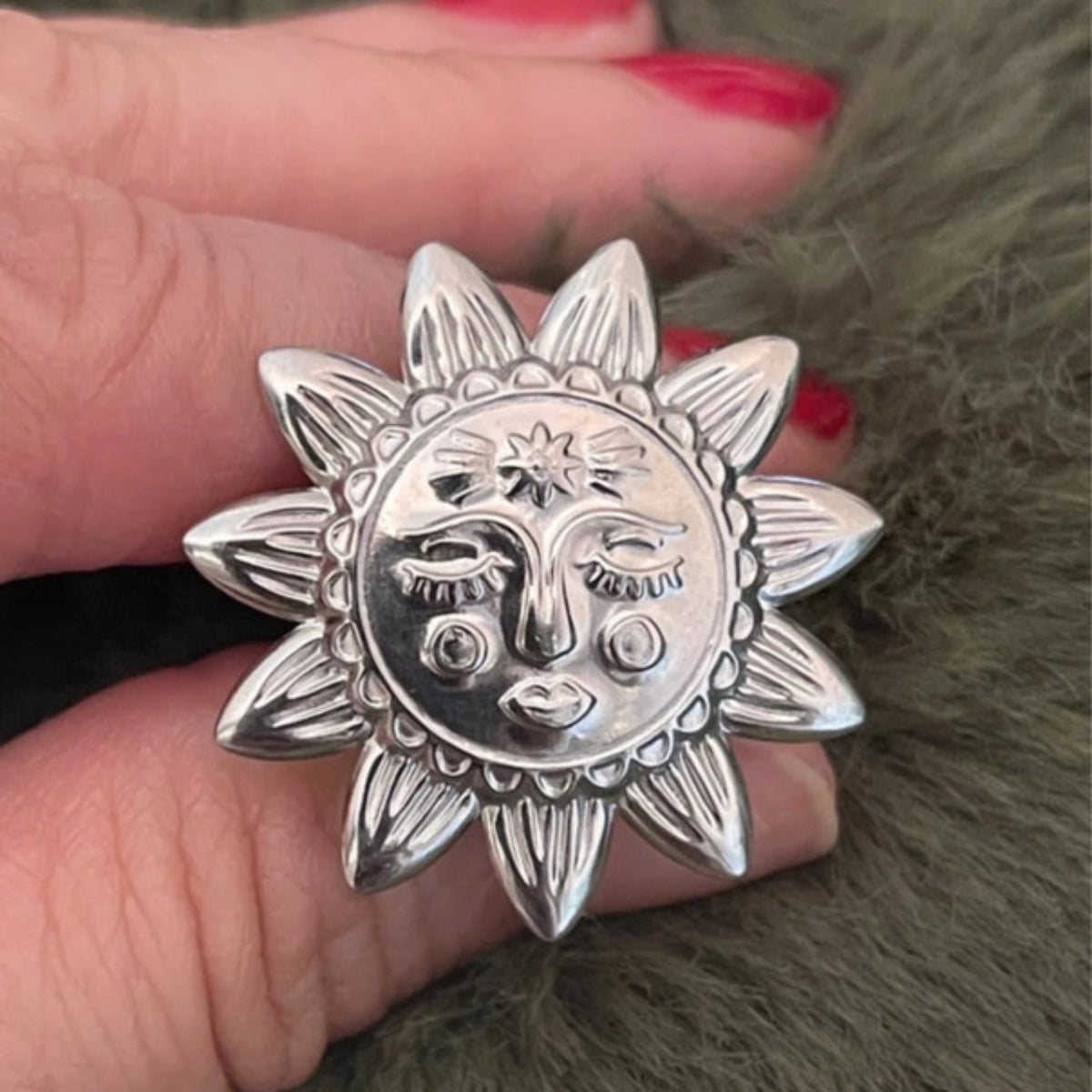 Silver sun-shaped ring with face design held between fingers on a textured surface.