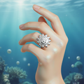 A silver sun-face ring worn on a hand, set against an underwater background with blue water and coral.