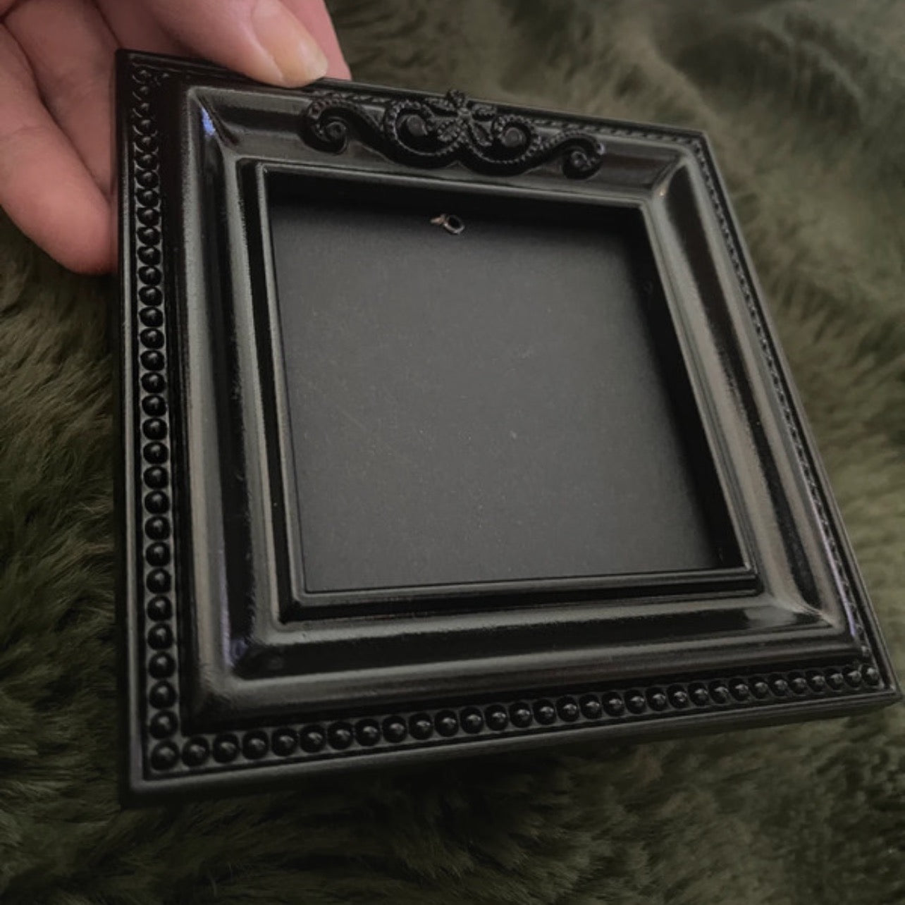 Black decorative picture frame held by a hand on a textured green surface.