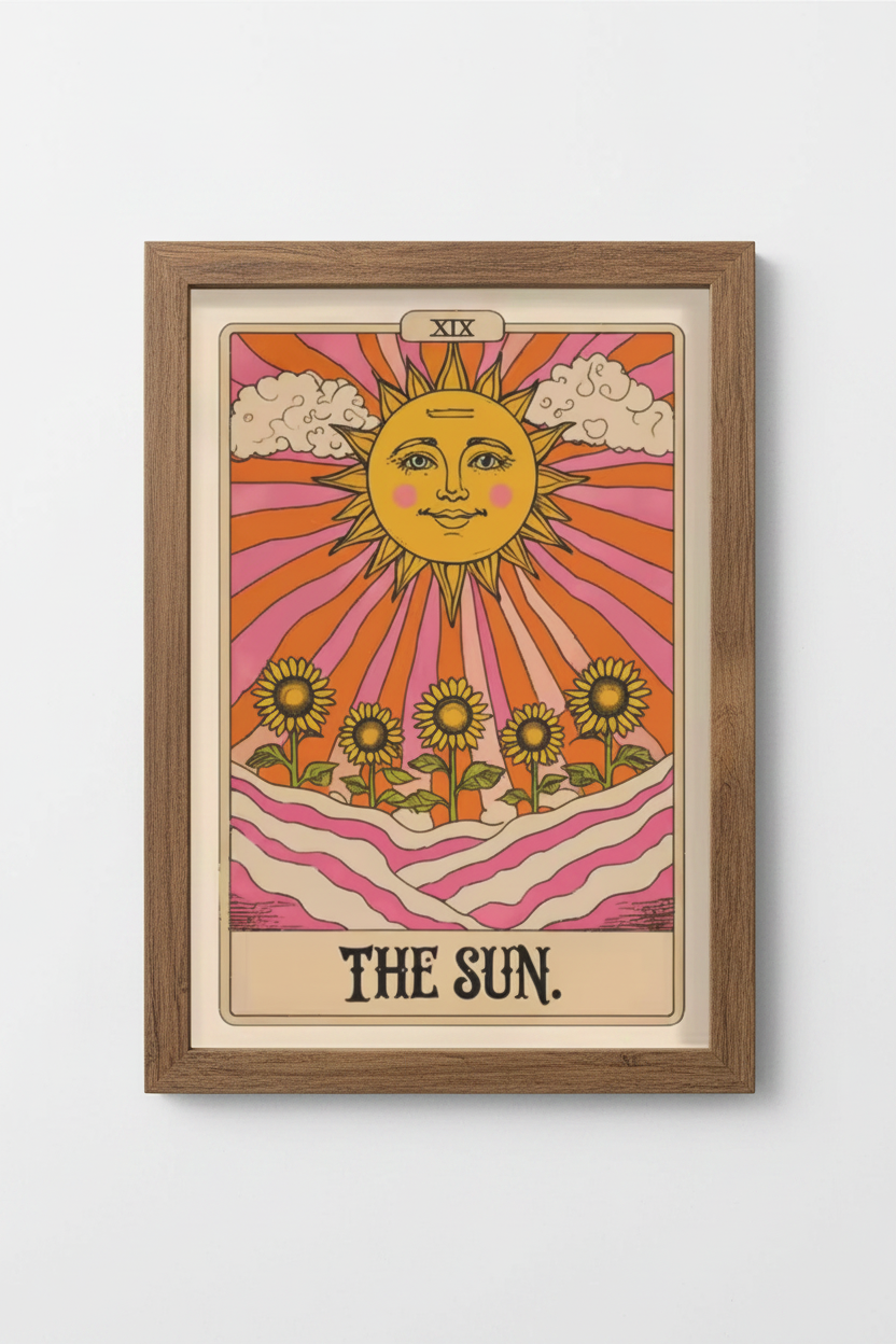 A framed art print of 'The Sun' tarot card, featuring a warm retro design with a smiling yellow sun, pink and orange rays, sunflowers, and wavy pink hills, set in a light brown wooden frame against a white wall.